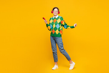 Fototapeta premium Full body young woman she wear glasses green sweater casual clothes listen to music in headphones use mobile cell phone isolated on plain orange yellow background studio portrait. Lifestyle concept.