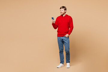 Fototapeta premium Full body young smiling happy man wear red hoodie casual clothes hold in hand use mobile cell phone typing message chat online isolated on pastel plain light beige background studio Lifestyle concept