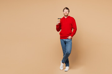 Full body young Caucasian man he wear red hoodie casual clothes hold takeaway delivery craft paper brown cup coffee to go isolated on pastel plain beige background studio portrait. Lifestyle concept.
