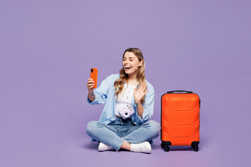 Traveler woman wear blue clothes hold use mobile cell phone waving hand sit isolated on plain purple background. Tourist travel abroad in free spare time rest getaway. Air flight trip journey concept.