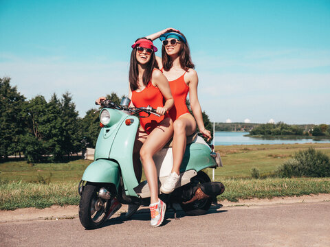 Fototapeta Two young beautiful smiling hipster female in summer red bathing suits. Sexy carefree women driving retro motorbike. Positive models having fun, riding classic Italian scooter. In swimwear