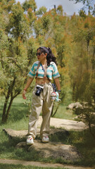 Fototapeta premium A young woman stands in a lush green landscape, wearing stylish casual attire, sunglasses, and holding a plush toy, embodying a relaxed outdoor vibe.