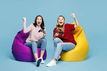 Full body winner young couple two friends women wear red casual clothes together sit in bag chair hold in hand play pc game with joystick console isolated on plain blue background. Lifestyle concept. © ViDi Studio