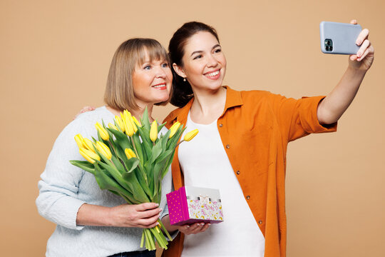 Elder parent mom 50s years old young daughter two women together wear casual clothes hold bouquet of tulips do selfie shot on mobile cell phone isolated on plain beige background. Family day concept.