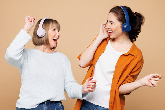 Elder cool parent mom 50s years old with young adult daughter two women together wear casual clothes listen to music in headphones isolated on plain pastel light beige background. Family day concept.