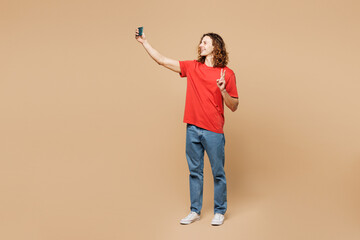 Full body young man he wears red t-shirt casual clothes doing selfie shot on mobile cell phone post photo on social network isolated on plain pastel light beige background studio. Lifestyle concept.