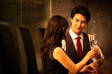 A well-dressed man and woman toast with champagne glasses while sitting at a modern upscale bar. The man wears a white shirt with a dark vest and red tie, and the woman is dressed in a black sleeveles