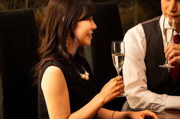 A man in a white shirt and vest and a woman in a black dress toast with champagne glasses at a high-end bar. The couple is smiling and enjoying a relaxed evening in a dimly lit, elegant setting.