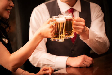 A close-up shot of a man and woman toasting with beer mugs in a warmly lit bar. The image focuses on their hands and glasses, emphasizing the golden color of the beer and the festive atmosphere.