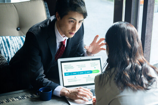 A businessman in formal attire is pointing to a laptop screen and explaining data to a female colleague across the table. They are seated by a bright window in a stylish café setting with a coffee mug