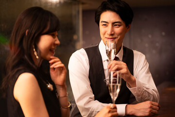 A man and woman dressed formally are smiling at each other while toasting with champagne glasses. They are seated across from each other at a wooden bar with warm ambient lighting.