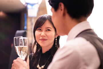 A woman in a sleeveless black dress smiles warmly at her partner while holding a champagne glass during a romantic evening at a high-end bar. Elegant jewelry and ambient lighting add to the luxurious 