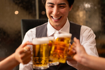 A cheerful man in formal attire smiles as he raises a glass of beer with friends. The warm lighting and joyful expression capture the spirit of celebration 