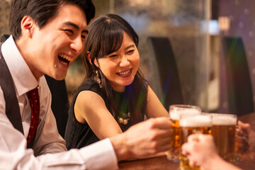 A joyful group of 4 friends toasts with beer mugs while sitting at a polished wooden bar. The...