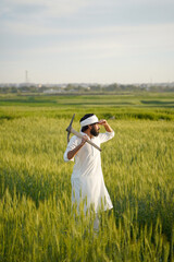 A Pakistani Indian farmer in traditional white attire carries a pickaxe on his shoulder while surveying a lush green wheat field.