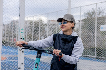 Woman riding a scooter near a fenced sports court in a suburban area