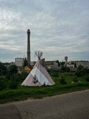 A conical tent, resembling a teepee, stands prominently in a grassy area.  In the background, a...