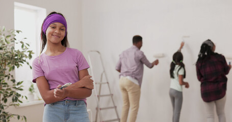 Obraz premium A smiling girl in a purple blouse and headband looks into the camera, holding a paintbrush, laughing. In the background, a family paints the walls next to a ladder during the apartment renovation.