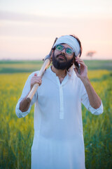 Naklejka premium A Pakistani Indian farmer wearing traditional white attire and a turban, holding a hoe on his shoulder while talking on a mobile phone.