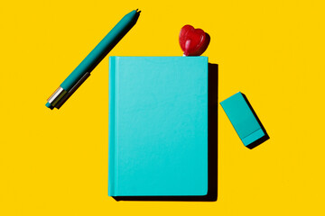 Turquoise notebook with pen, eraser, and heart-shaped lollipop on yellow background