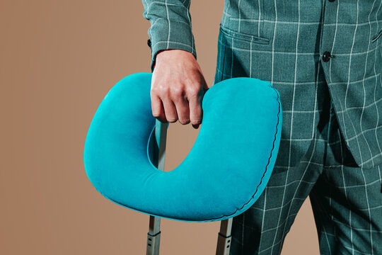 Businessman in gray suit holding a blue inflatable travel pillow with a trolley suitcase against a brown background