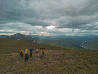 A group of travelers in bright clothes stand on the peaks of the mountain during the golden hour, the rays of the sun break through the clouds and fall on a green meadow.