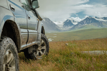 An off-road tourist car for traveling stands in a field in a meadow against the background of snowy mountains on off-road mud tires. Off-road tires on the background of mountains.