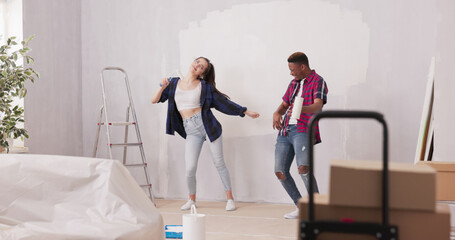Fototapeta premium Smiling, cheerful friends have fun renovating their apartment, playfully painting walls with rollers. The room is filled with laughter, with furniture securely covered to protect against dirt.
