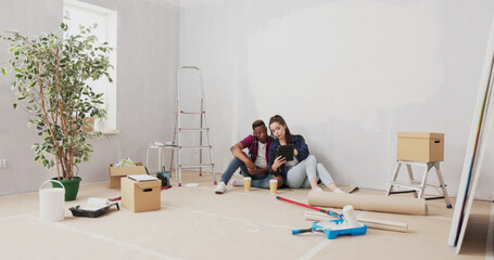 A loving couple relaxes on the floor of their newly renovated apartment, drinking take-out coffee and scrolling through a tablet for furniture and decoration ideas.
