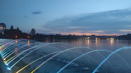 Illuminated water fountains gracefully arc over a tranquil river at dusk, reflecting vibrant city...