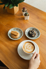 A latte with foam art with a hand holding the coffee cup and pastries on a wooden table