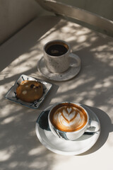 A latte with heart-shaped foam art and black coffee, paired with a chocolate chip cookie on a sunlit on white table.