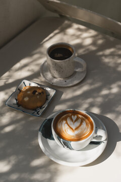 A latte with heart-shaped foam art and black coffee, paired with a chocolate chip cookie on a sunlit on white table.