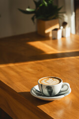A latte with foam art on a wooden table, bathed in afternoon sunlight with a potted plant adding charm.