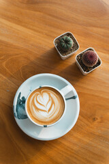 A latte with foam art on a wooden table, bathed in afternoon sunlight with a potted plant adding charm.
