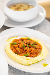 Plate with creamy mashed potatoes topped with savory meat stew and fresh dill garnish, accompanied by a bowl of soup on a white saucer. Close-up presentation in a cozy dining setting