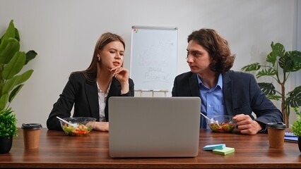 Business team of man and woman sitting at desk in the office having dinner break with food delivery, people talking working on laptop eating healthy salads.