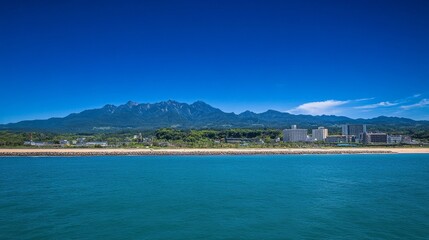 Scenic coastal landscape with bright blue sky, sunlit mountains, and shimmering sea reflecting sunlight. Clear horizon with distant seaside buildings enhancing the picturesque view.
