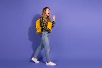 Young woman in casual attire with a backpack waving cheerfully against a purple background
