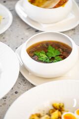 A white bowl filled with dark broth garnished with fresh parsley, placed on a plate with bread, potatoes, and eggs in the background, offering a homemade meal vibe