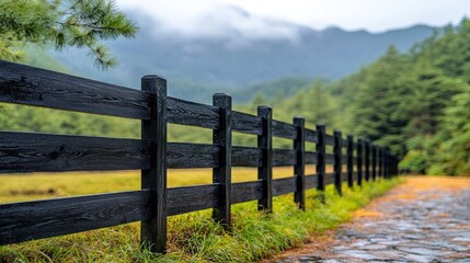 Fototapeta premium Dark wooden fence lines a grassy path towards misty mountains
