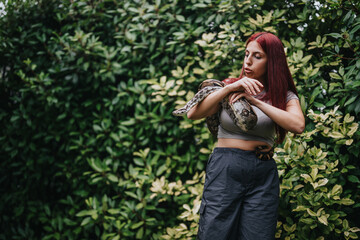 A young girl with red hair holds a snake confidently while surrounded by lush green foliage, demonstrating bravery and a connection with nature.