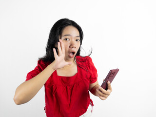Naklejka premium A young beautiful Asian woman wearing red t-shirt and holding her phone is shouting and screaming loud with a hand on her mouth, isolated by white background.
