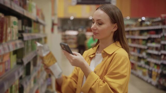 In a grocery store, a woman scans a coffee package with her phone while browsing the aisles. She appears focused on her shopping task, selecting products carefully.