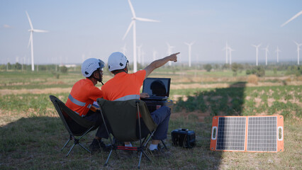 At a wind farm, two Asian inspectors are reviewing laptop data to analyze system performance and blade rotation, with power supplied by a portable solar panel. Engineer Wind Turbine.