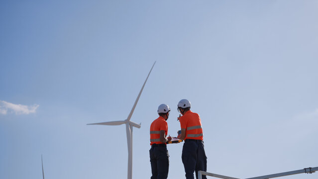 In a wind farm, two Asian inspectors are reviewing data on a tablet and clipboard to analyze system performance and blade rotation at varying wind speeds. Engineer Wind Turbine.