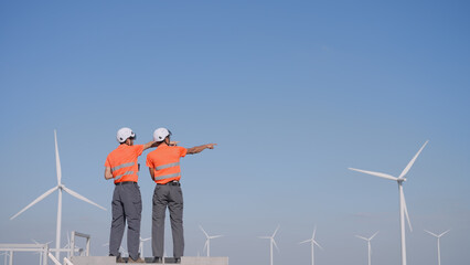 In a wind farm, a rear view shows two Asian inspectors are reviewing data on a tablet and clipboard to analyze system performance and blade rotation at varying wind speeds. Engineer Wind Turbine.