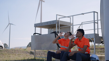 Two wind energy technicians in reflective orange uniforms drink water while seated near turbine, taking a break under sunny skies in a renewable energy field.