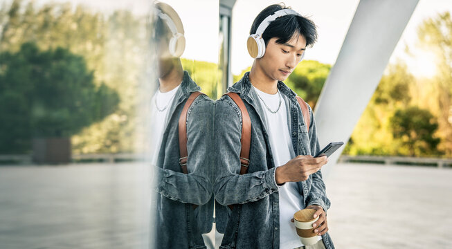 Millenial asian man wearing headphones using smart mobile phone device outdoors - Young male person listening podcast with smartphone - Technolohy life style concept - Powered by Adobe