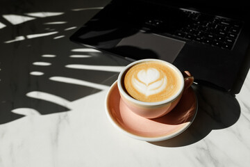 metallic laptop and pink cup with cappuccino coffee on white table at sunny work day